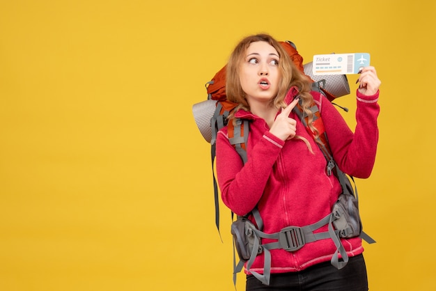 Vista frontal de uma jovem garota viajando surpresa com máscara médica, segurando e apontando o bilhete