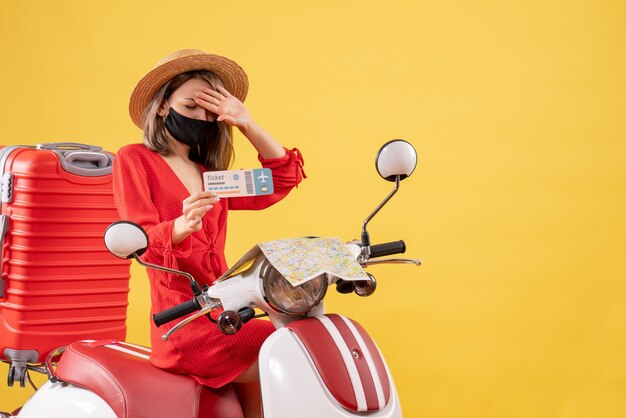 Vista frontal de uma jovem insatisfeita em uma motocicleta com uma mala vermelha segurando o bilhete