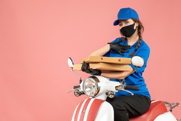 Vista frontal de uma jovem mensageira insegura usando máscara médica e luvas, sentada na scooter, entregando pedidos em fundo de pêssego pastel