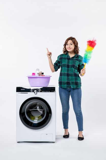 Vista frontal de uma jovem mulher com a máquina de lavar roupa segurando o limpador de pó na parede branca