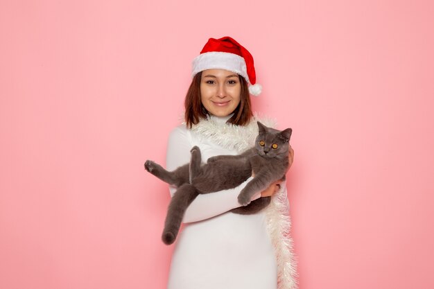 Vista frontal de uma jovem mulher com boné vermelho segurando um gatinho fofo cinza na parede rosa