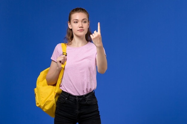 Vista frontal de uma jovem mulher com camiseta rosa e mochila amarela na parede azul claro