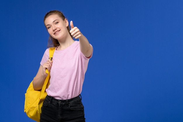 Foto grátis vista frontal de uma jovem mulher com uma camiseta rosa e uma mochila amarela posando sorrindo na parede azul clara