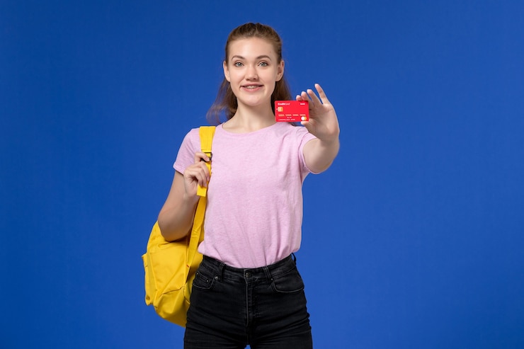 Foto grátis vista frontal de uma jovem mulher com uma camiseta rosa e uma mochila amarela segurando um cartão de plástico vermelho na parede azul