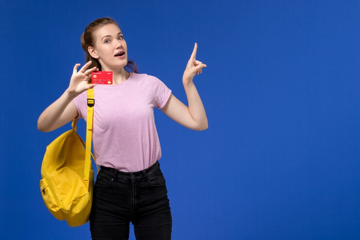 Foto grátis vista frontal de uma jovem mulher com uma camiseta rosa e uma mochila amarela segurando um cartão de plástico vermelho na parede azul