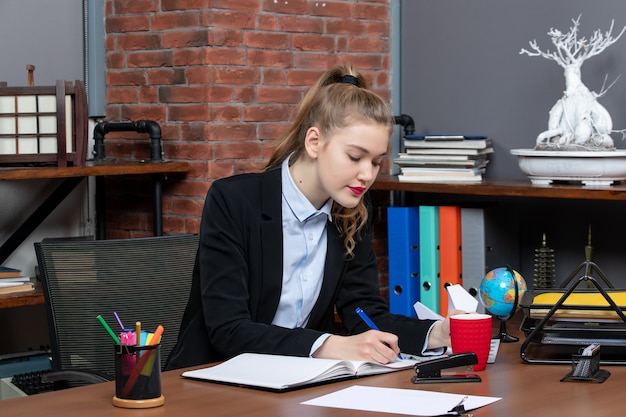 Vista frontal de uma jovem mulher concentrada, sentada à mesa e escrevendo em um documento no escritório