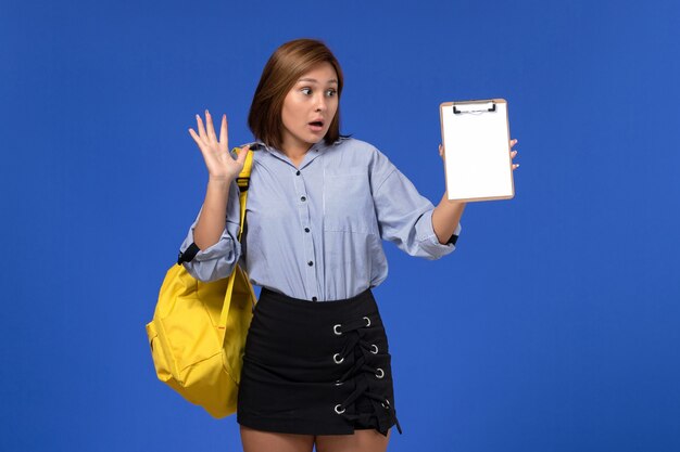 Vista frontal de uma jovem mulher de camisa azul com saia preta, mochila amarela e segurando o bloco de notas na parede azul claro