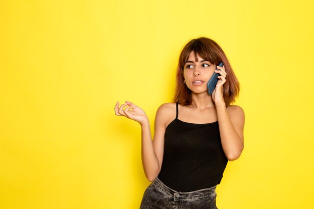 Vista frontal de uma jovem mulher de camisa preta e calça jeans cinza falando ao telefone na parede amarela