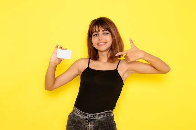 Vista frontal de uma jovem mulher de camisa preta e calça jeans cinza segurando um cartão de plástico na parede amarela