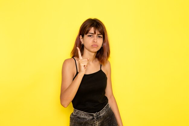 Vista frontal de uma jovem mulher de camisa preta e jeans cinza, ameaçando com o dedo na parede amarela