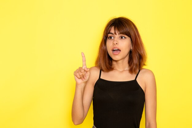 Vista frontal de uma jovem mulher de camisa preta levantando o dedo na parede amarelo-claro