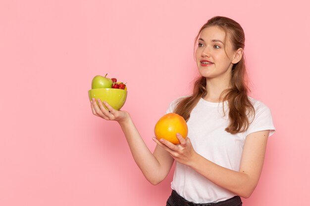 Vista frontal de uma jovem mulher de camiseta branca segurando um prato com frutas frescas e toranja na parede rosa