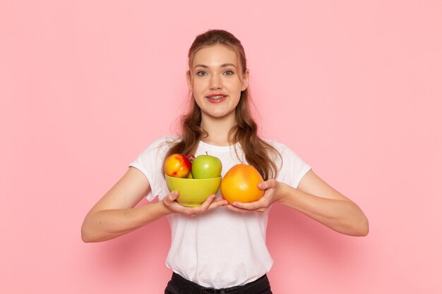 Vista frontal de uma jovem mulher de camiseta branca segurando um prato com frutas frescas e toranja na parede rosa