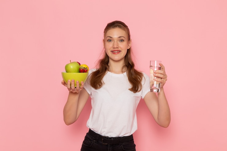 Foto grátis vista frontal de uma jovem mulher em uma camiseta branca segurando um prato com frutas e um copo de água na parede rosa