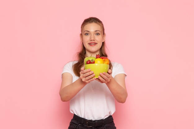 Vista frontal de uma jovem mulher em uma camiseta branca segurando um prato com frutas frescas na parede rosa claro