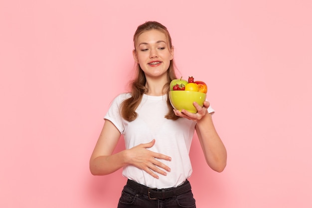 Vista frontal de uma jovem mulher em uma camiseta branca segurando um prato com frutas frescas na parede rosa claro