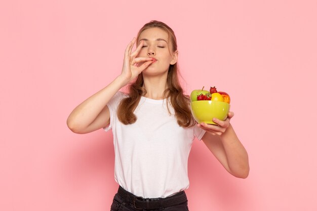 Vista frontal de uma jovem mulher em uma camiseta branca segurando um prato com frutas frescas na parede rosa