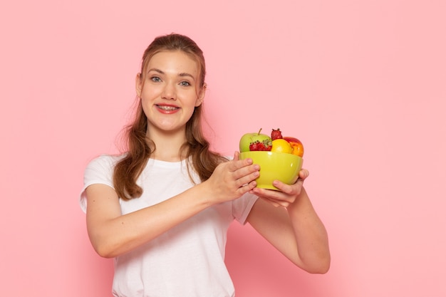 Vista frontal de uma jovem mulher em uma camiseta branca segurando um prato com frutas frescas, sorrindo na parede rosa claro