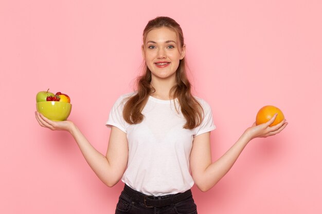 Vista frontal de uma jovem mulher em uma camiseta branca segurando um prato com frutas frescas sorrindo