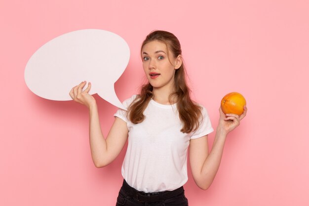 Vista frontal de uma jovem mulher em uma camiseta branca segurando uma grapefruit e uma placa branca na parede rosa