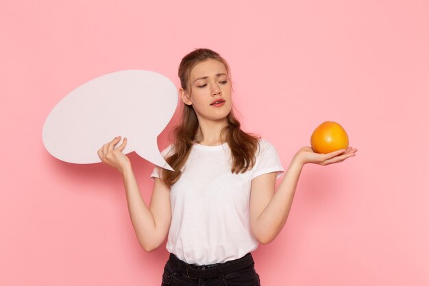 Vista frontal de uma jovem mulher em uma camiseta branca segurando uma grapefruit e uma placa branca na parede rosa