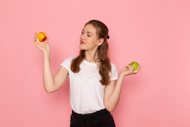 Vista frontal de uma jovem mulher em uma camiseta branca segurando uma maçã verde fresca com pêssego na parede rosa claro