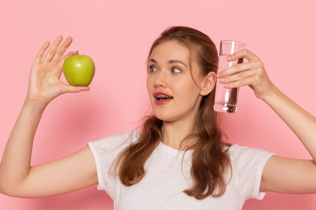 Vista frontal de uma jovem mulher em uma camiseta branca segurando uma maçã verde fresca e um copo de água na parede rosa