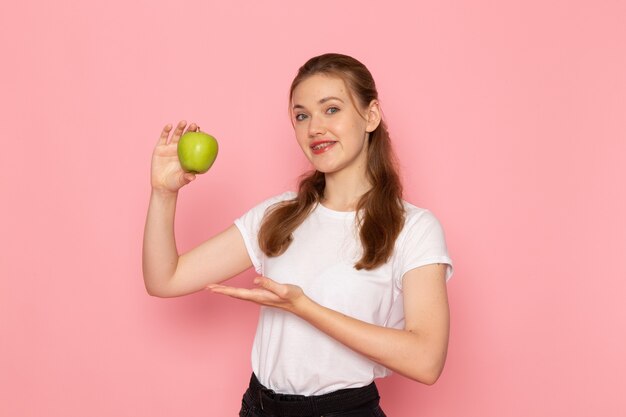 Vista frontal de uma jovem mulher em uma camiseta branca segurando uma maçã verde sorrindo na parede rosa claro