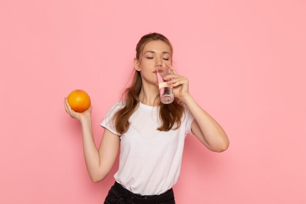 Vista frontal de uma jovem mulher em uma camiseta branca segurando uma toranja fresca e um copo de água bebendo na parede rosa