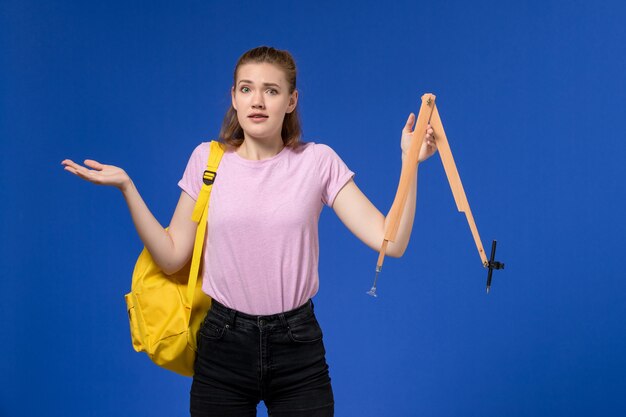 Vista frontal de uma jovem mulher em uma camiseta rosa, usando uma mochila amarela, segurando uma figura de madeira na parede azul claro