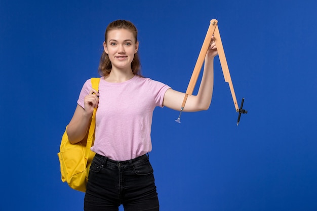 Foto grátis vista frontal de uma jovem mulher em uma camiseta rosa, usando uma mochila amarela, segurando uma figura de madeira na parede azul