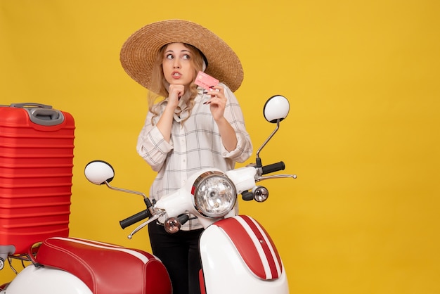 Vista frontal de uma jovem pensativa com chapéu, recolhendo a bagagem, sentada na motocicleta e segurando o cartão do banco
