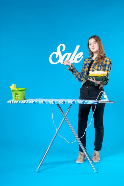 Vista frontal de uma jovem segurando o ícone de venda de roupas dobradas e em pé atrás da tábua de passar na lavanderia