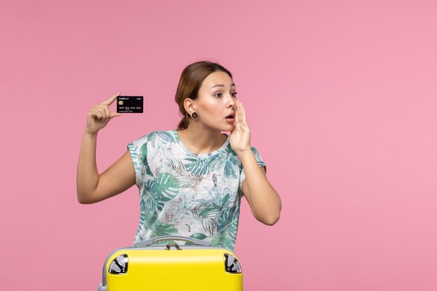 Vista frontal de uma jovem segurando um cartão de banco preto chamando alguém na parede rosa