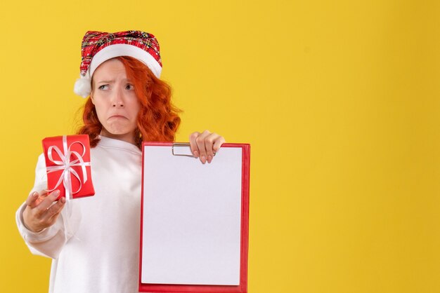 Vista frontal de uma jovem segurando um pequeno presente de Natal e uma nota de arquivo com uma cara triste na parede amarela