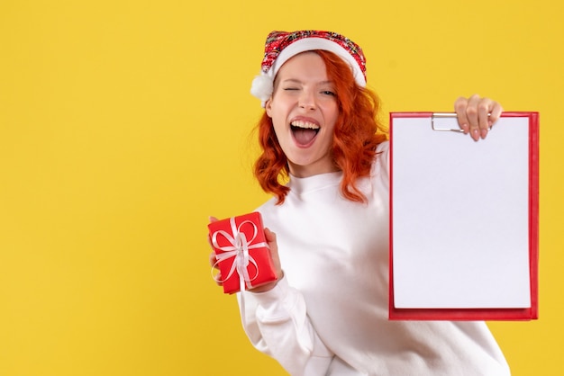 Vista frontal de uma jovem segurando um pequeno presente de Natal e uma nota de arquivo na parede amarela
