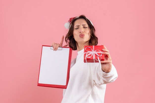 Vista frontal de uma jovem segurando um pequeno presente de Natal e uma nota de arquivo na parede rosa