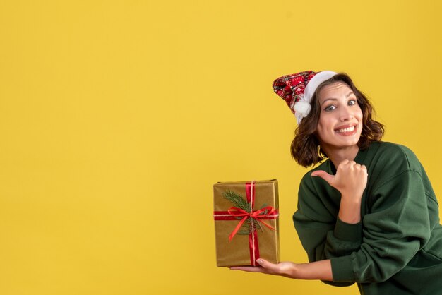 Vista frontal de uma jovem segurando um pequeno presente de Natal na parede amarela