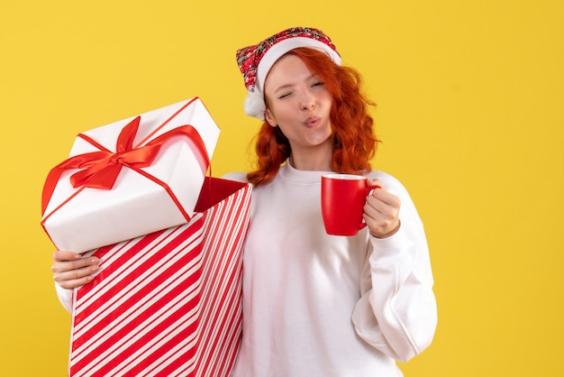 Vista frontal de uma jovem segurando um presente de natal e uma xícara de chá na parede amarela