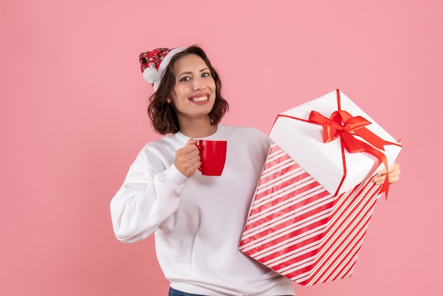 Vista frontal de uma jovem segurando um presente de Natal e uma xícara de chá na parede rosa