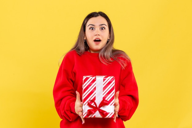 Vista frontal de uma jovem segurando um presente de natal na parede amarela