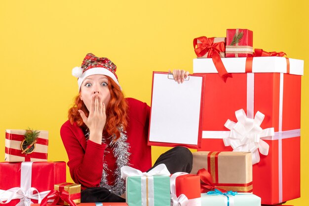 Vista frontal de uma jovem sentada ao redor de diferentes presentes de Natal na parede amarela