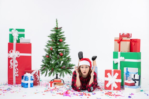 Vista frontal de uma jovem sentada ao redor de presentes de Natal e uma pequena árvore de férias na parede branca
