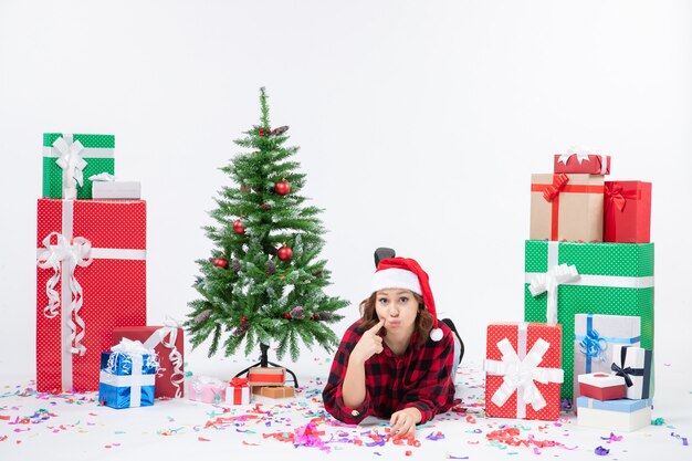Vista frontal de uma jovem sentada ao redor de presentes de Natal e uma pequena árvore de férias na parede branca