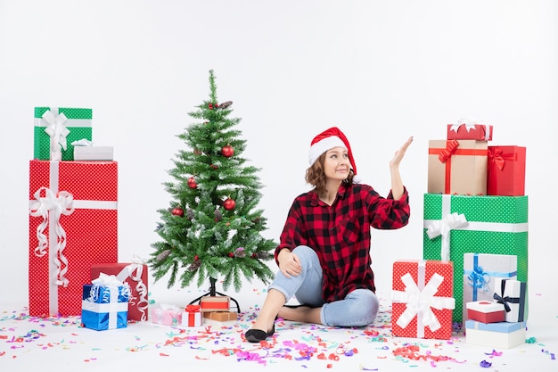 Vista frontal de uma jovem sentada ao redor de presentes de Natal e uma pequena árvore de férias na parede branca