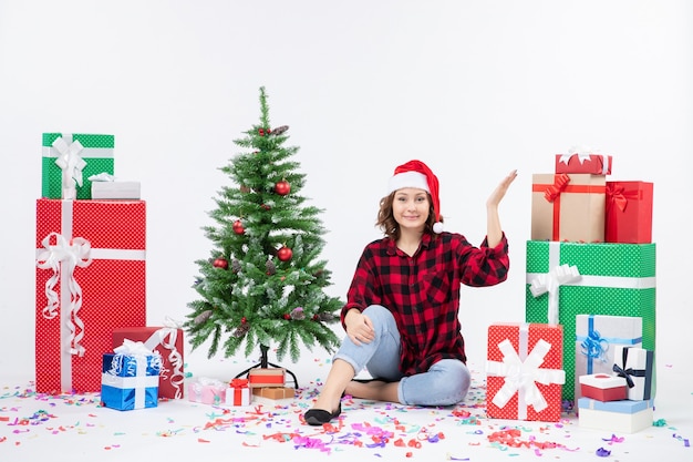 Vista frontal de uma jovem sentada ao redor de presentes de natal e uma pequena árvore de férias na parede branca