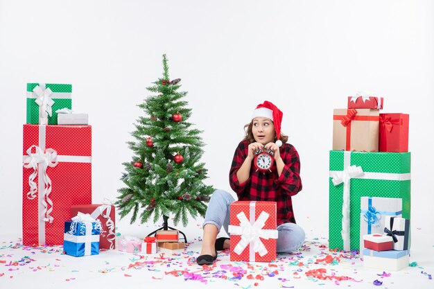 Vista frontal de uma jovem sentada ao redor de presentes de Natal segurando relógios na parede branca