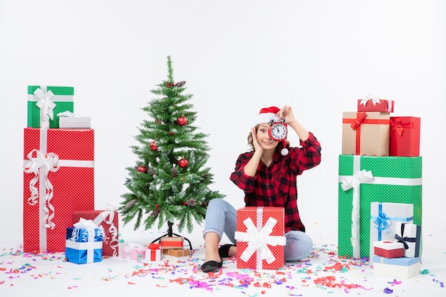 Vista frontal de uma jovem sentada ao redor de presentes de Natal segurando relógios na parede branca
