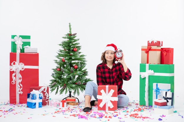 Vista frontal de uma jovem sentada ao redor de presentes de Natal segurando relógios na parede branca