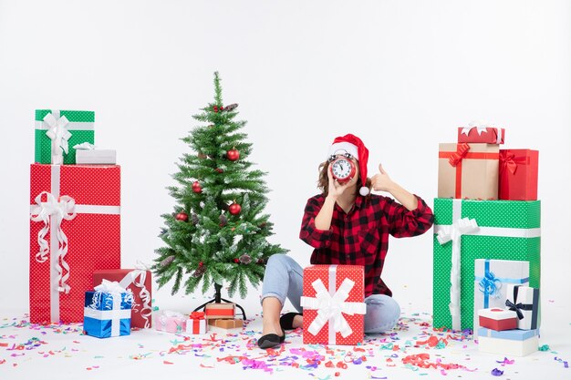 Vista frontal de uma jovem sentada ao redor de presentes de Natal segurando relógios na parede branca